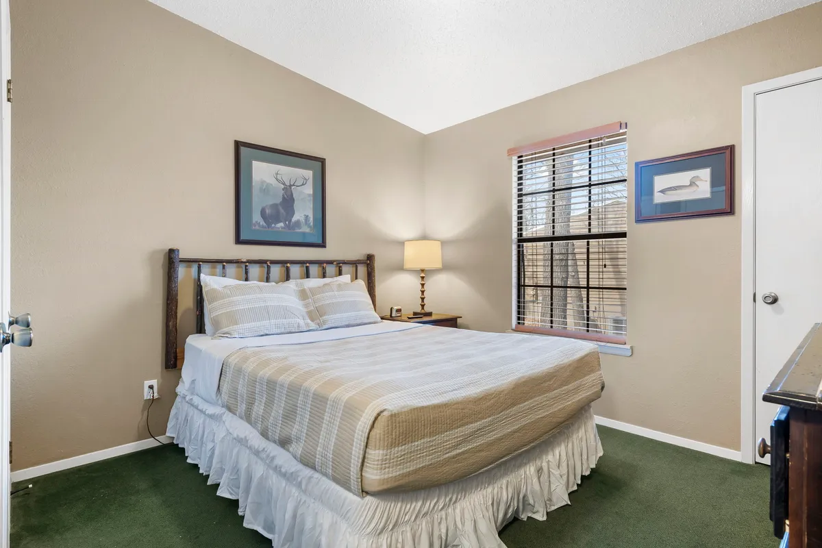 An inviting bedroom with a comfortable bed, a bedside lamp, a large window letting in morning light, and a piece of colorful artwork adorning the wall. The room feels bright, calm, and ideal for relaxation or a quiet morning start.