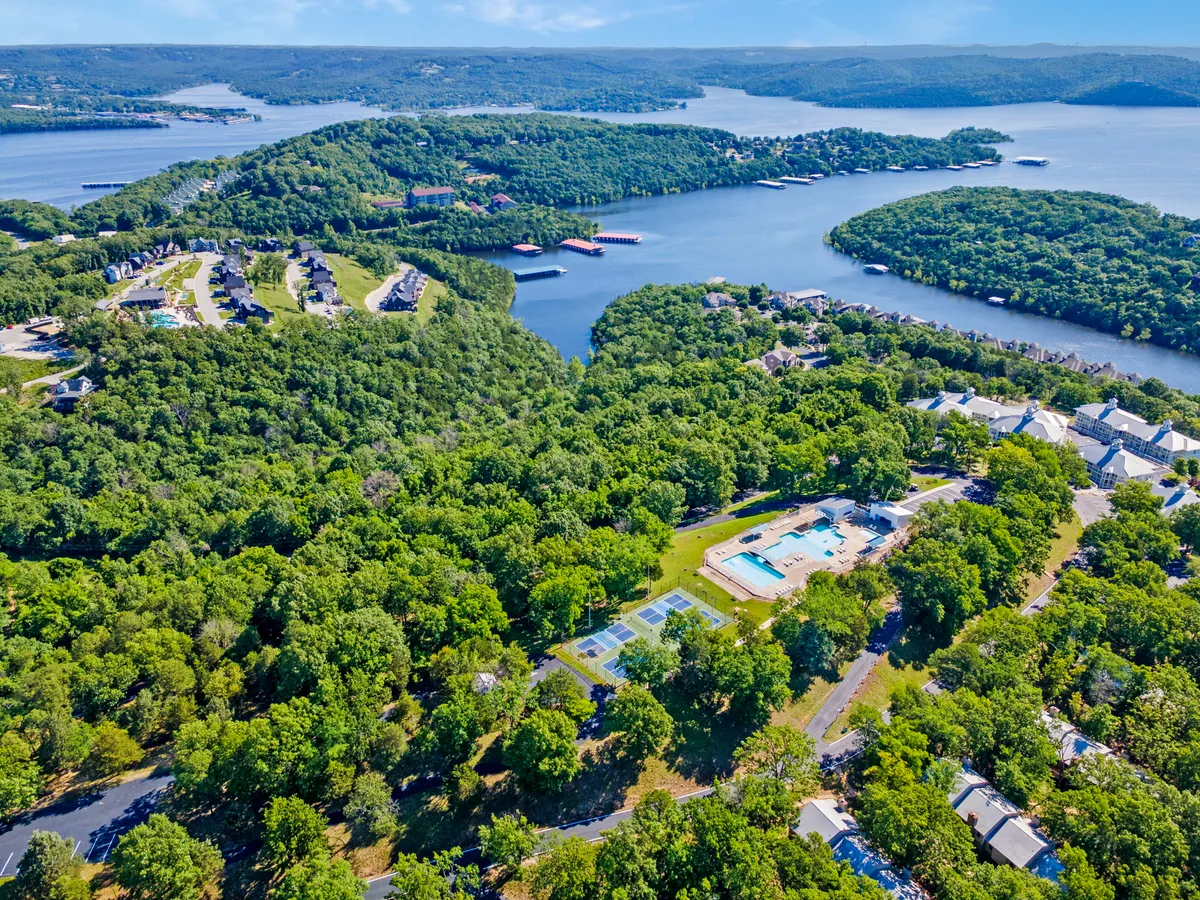 A vibrant resort view featuring a sparkling swimming pool, adjacent tennis courts, lush landscaping, waterfront access, and residential buildings in the background. Guests can be seen enjoying sun-soaked days by the pool and engaging in outdoor activities like boating and hiking.