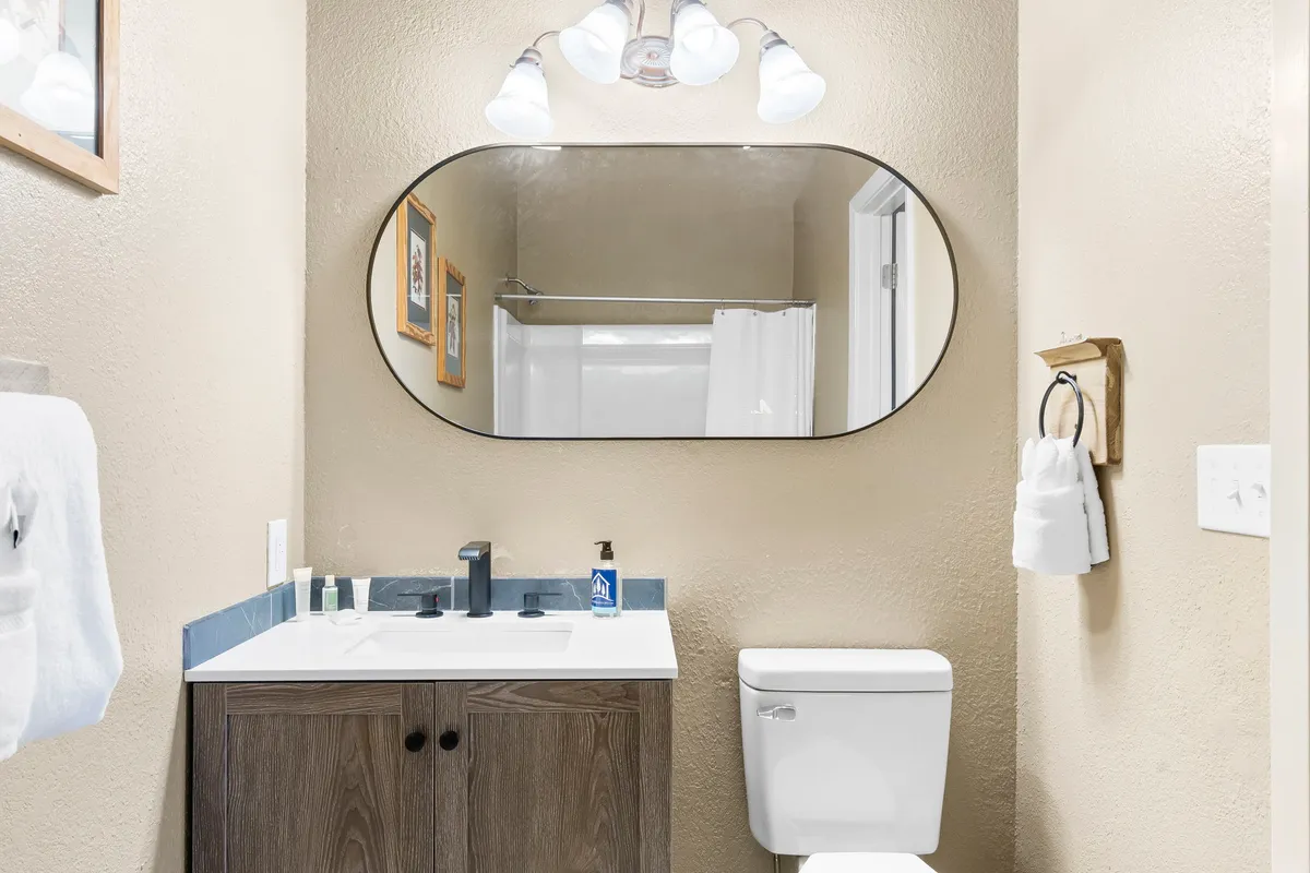 A bright, functional bathroom with an oval mirror above the sink, modern fixtures, a light fixture providing soft illumination, a toilet, and a piece of wall art. The space is tidy and serene, promoting relaxation and personal care.