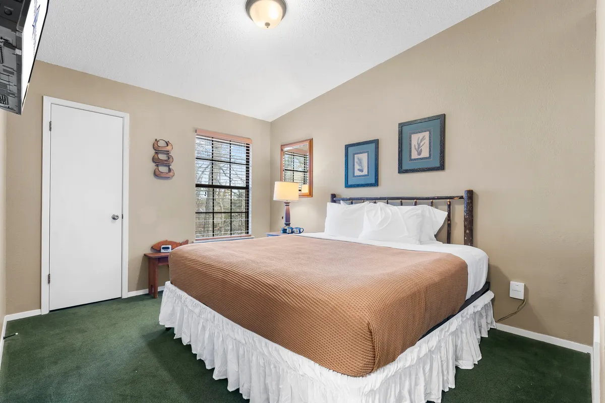 A cozy bedroom featuring a bed with a warm brown coverlet, soft lighting from a bedside lamp, framed artwork on the walls, a window with blinds letting in filtered natural light, and a small nightstand. The ambiance is peaceful and inviting, emphasizing relaxation and comfort.