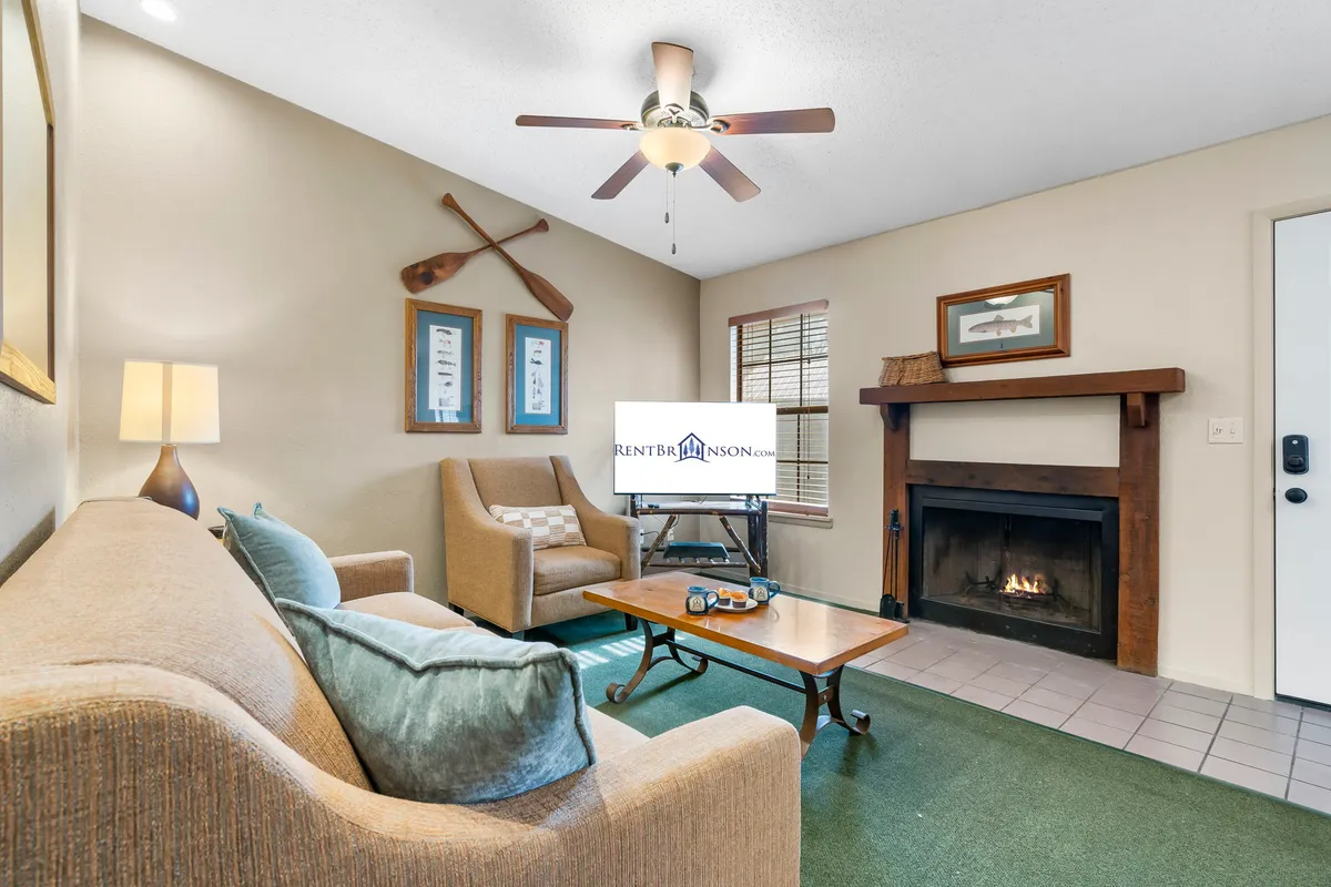The image showcases a cozy interior living area featuring a glowing stone fireplace, comfortable seating, warm lighting from a lamp, and charming decorative oars on the wall. The inviting space is perfect for relaxing, reading, or gathering for board games with family and friends.
