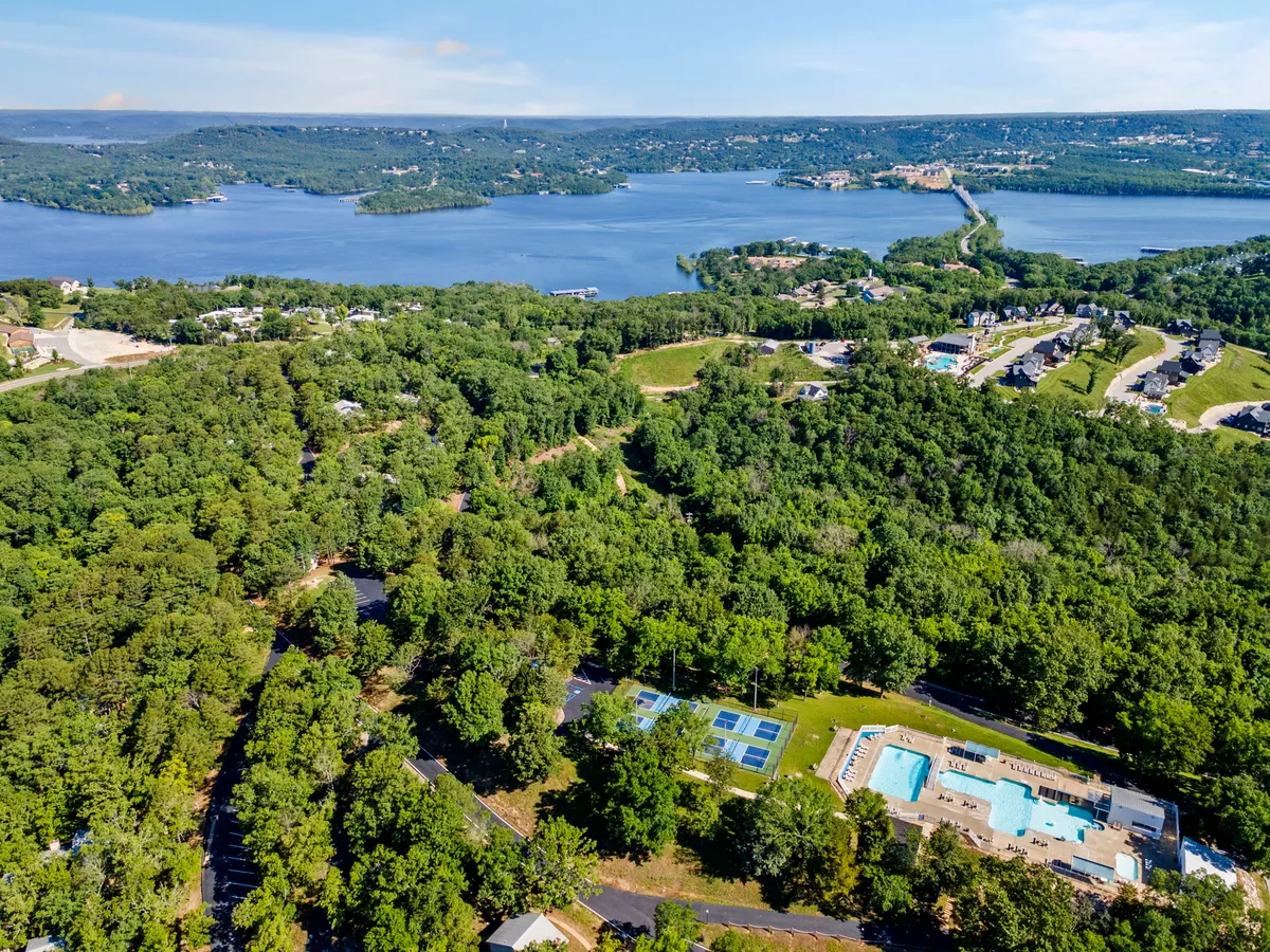 A broad, panoramic view encompassing a scenic lake, swimming pool, tennis courts, lush greenery, and nearby residential areas. The image captures the scale and diversity of resort amenities amid the beautiful Ozark landscape, with nature trails and water activities suggested by the setting.