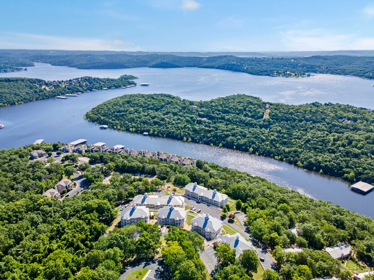 A tranquil river scene surrounded by lush greenery, with a small cluster of buildings nestled along the shoreline. The scene suggests opportunities for boating, hiking, fishing, and enjoying picnics by the water. The peaceful ambiance is perfect for watching sunsets and exploring the natural beauty of the Ozarks.
