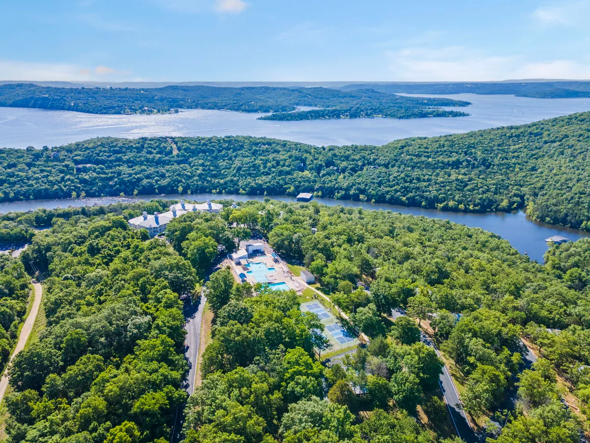 A wide exterior view showcasing lush greenery, a swimming pool, tennis courts, and a shimmering river and lake in the background. The scene is set during sunset, with the golden light highlighting walking trails surrounded by trees and nature.
